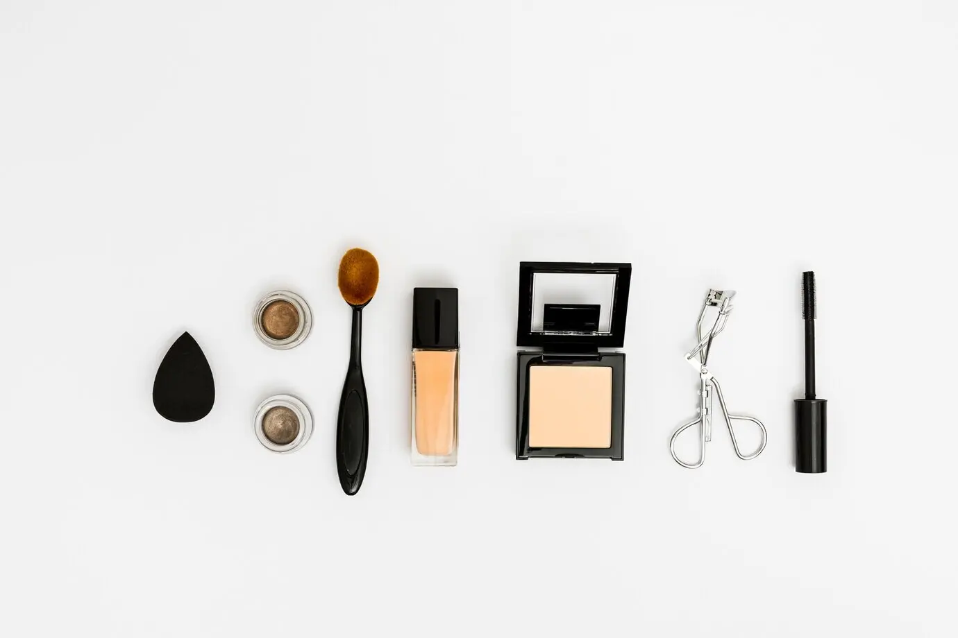 Draufsicht auf Make-up-Schwamm, Lidschatten, ovalen Pinsel, Kompaktpuder, Wimpernzange und Mascara-Bürste auf weißem Hintergrund.